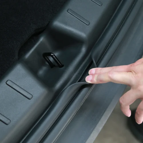 Close-up of a car trunk interior: a person pulls back the rubber edge seal near a small black tie-down hook in the trunk lip.