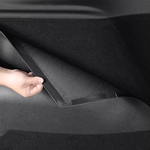 Close-up of a hand pulling a fabric strap along a car's floor panel, likely opening a hidden storage area under the trunk/cargo floor.