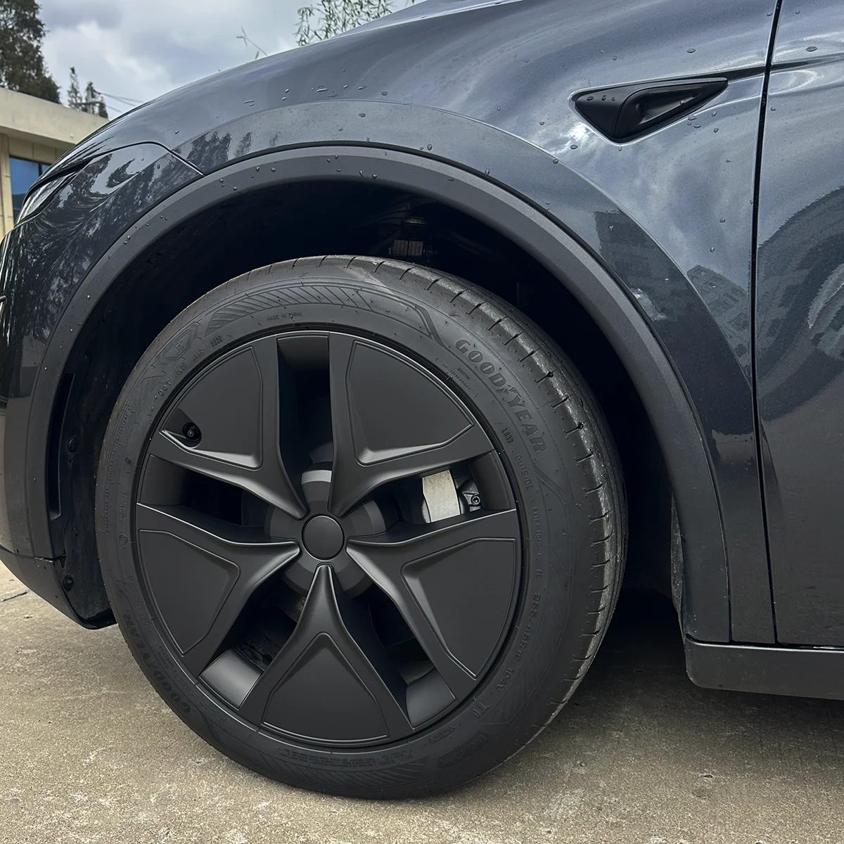 Close-up of a dark gray car wheel with an aerodynamic five-spoke cover, tire labeled Goodyear, and surrounding fender.