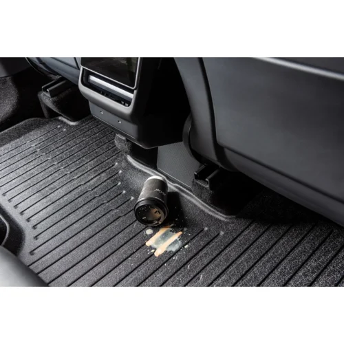 Camera lens sits on a car floor mat near the rear of the center console with a spilled liquid nearby