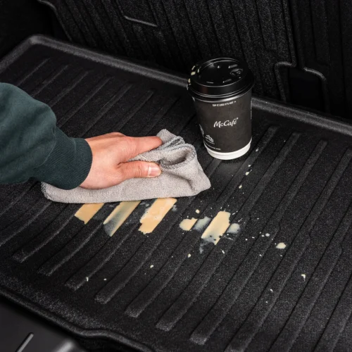 Person wiping a coffee spill from a black car trunk mat with a gray cloth and a McCafé cup nearby