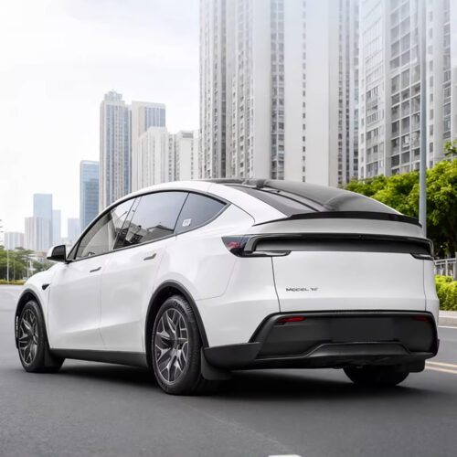 Rear three-quarter view of a white Model Y SUV driving on a city street with high-rise buildings in the background.